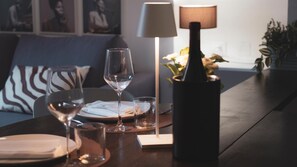 Une table à manger dressée avec des assiettes, des verres et une bouteille, avec une lampe et un vase de fleurs en arrière-plan.