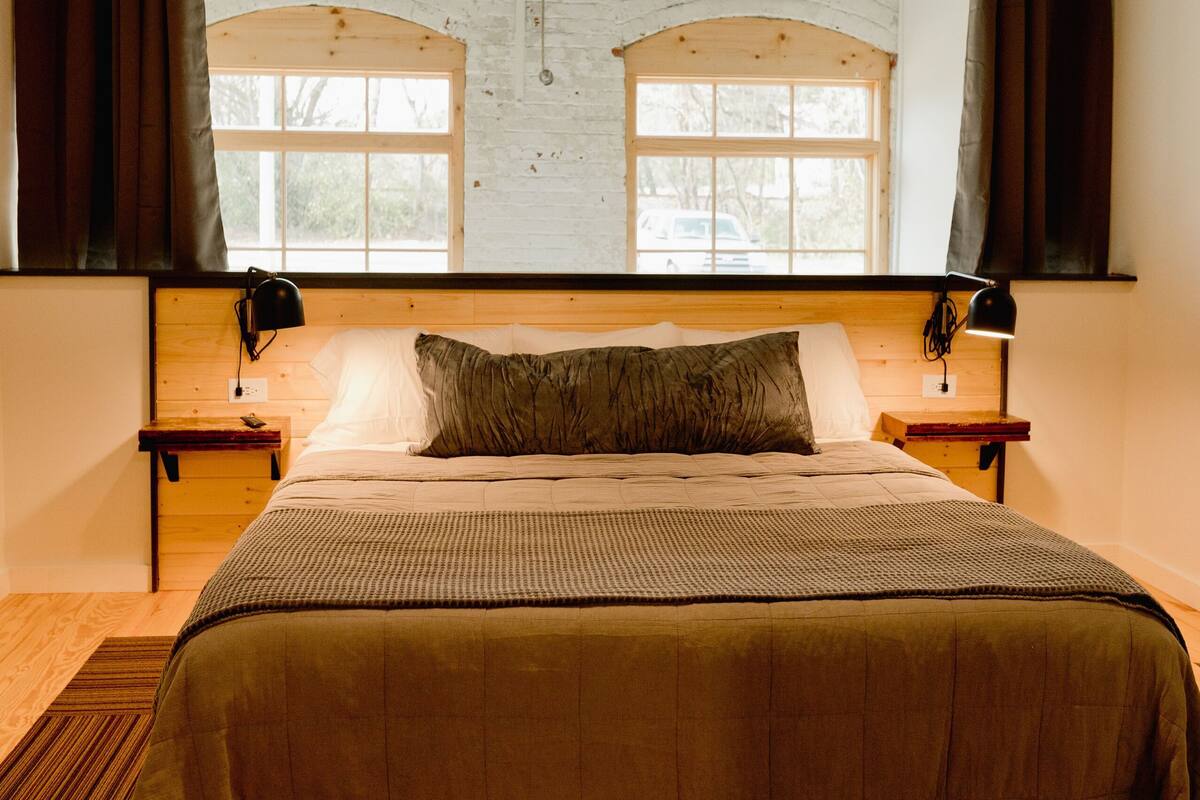 A bedroom with a large bed, wooden headboard, two bedside tables, a ceiling fan, and a view of the outside through arched windows.
