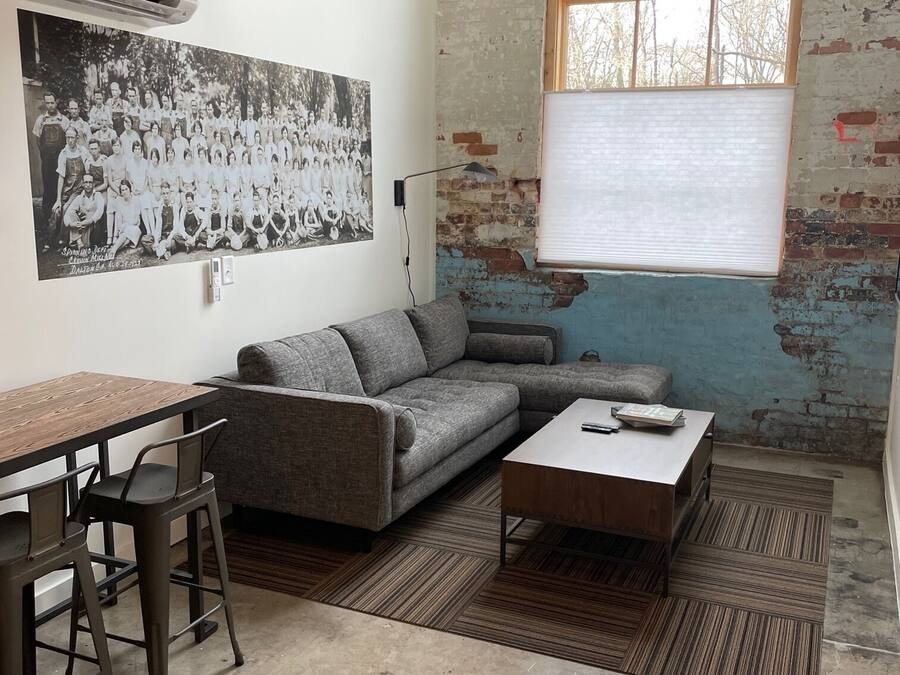 Una sala de estar con un sofá gris, una mesa de centro de madera y una zona de comedor con una mesa de madera y sillas de metal. Hay una fotografía grande en blanco y negro en la pared y una ventana con persianas.