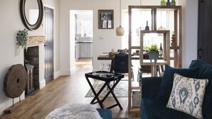 Een woonkamer met een donkerblauwe bank, een ronde salontafel en een zacht tapijt. Er is een open haard met een spiegel erboven, en een eethoek met een houten tafel en stoelen.