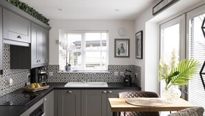 Een moderne keuken met grijze kastjes, een zwart aanrecht en een houten vloer. Er is een eethoek met een tafel en stoelen.