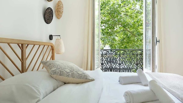 A bedroom with a wooden headboard, a bed with white linens, a patterned pillow, and a view of a balcony with greenery outside. - lovely apartment center of Paris (Paris)