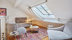 Ein gemütliches Schlafzimmer im Loft-Stil mit einem Oberlicht, einem gemusterten Sessel, einem grauen Sofa und einem kleinen Tisch mit einer Lampe.