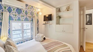 Ein Hotelzimmer mit Bett, Fenster mit gemusterten Vorhängen, einem an der Wand montierten Fernseher und einem Regal mit Dekoration.