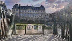 Une entrée fermée par un portail mène à un bâtiment historique. Un panneau indique qu’il est interdit d’y entrer avec des chiens, et une affiche appelle à la lutte contre le racisme.