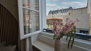 Une pièce avec une fenêtre donnant sur un paysage urbain, un banc en bois et un vase rempli de fleurs.