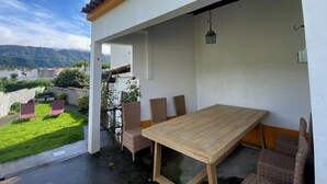 An outdoor patio with a wooden table and wicker chairs, overlooking a lush garden and distant hills.