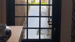 A view through a window with a grid pattern, leading to a courtyard with a wooden fence, a red chair, and greenery.