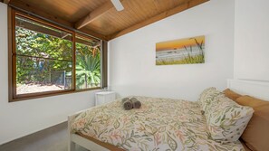 A bedroom with a bed, a window with a view of greenery, and a painting of a sunset.