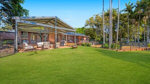 A covered outdoor seating area with a barbecue grill, surrounded by a well-maintained lawn and a fenced pool area.