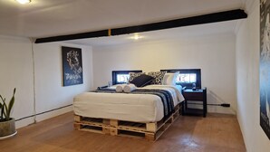 Ein Schlafzimmer mit einem Bett auf einer Holzpalette, einem Nachttisch mit Lampe, einem Wandbild und einer Topfpflanze.