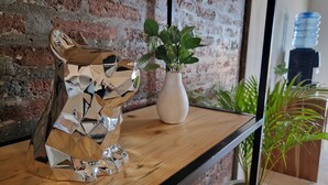 Eine Metallskulptur auf einem Holzregal, eine Vase mit Grünpflanzen und ein Wasserspender im Hintergrund.
