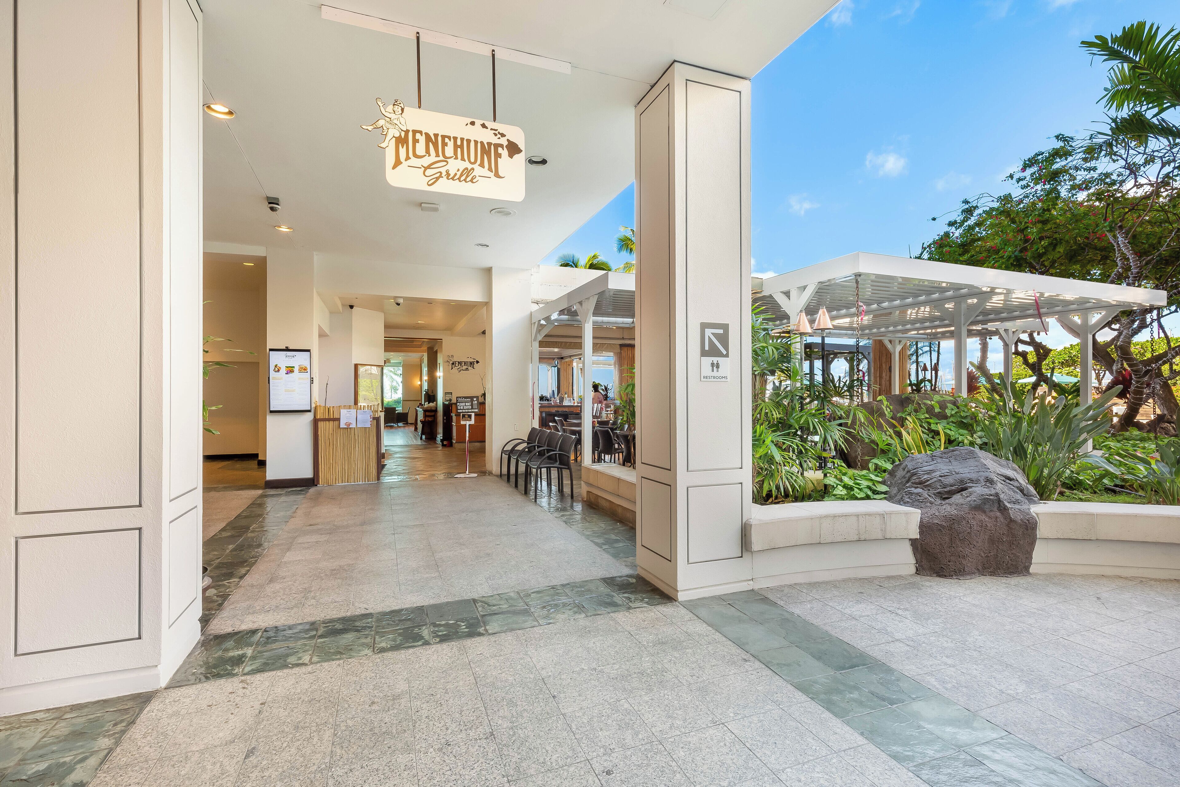 En modern hotellobby med en receptionsdisk, en sittgrupp och ett takförsett utomhusområde med växter.