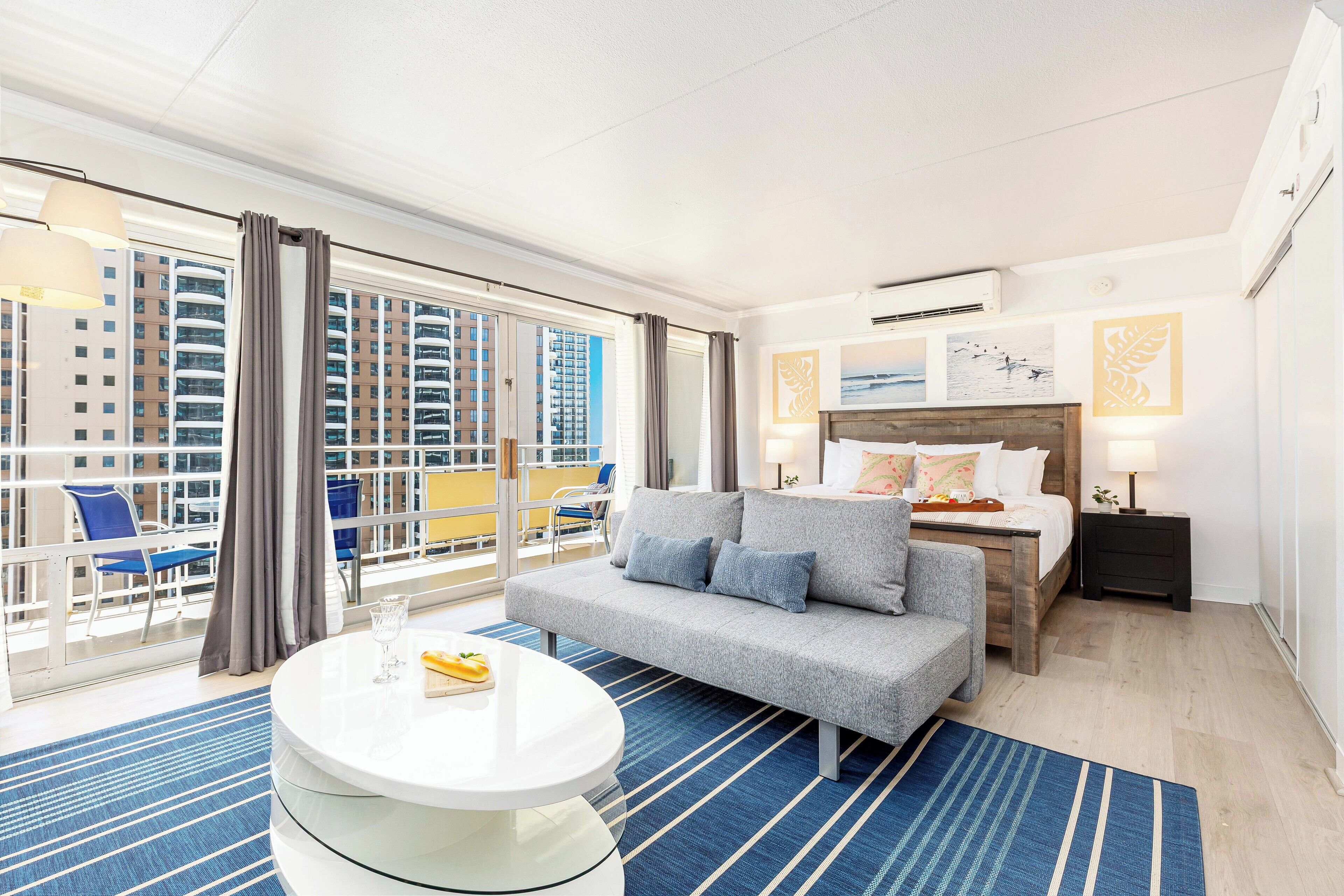 Een hotelkamer met een groot raam, een bank, een bed, een nachtkastje en een ronde tafel.