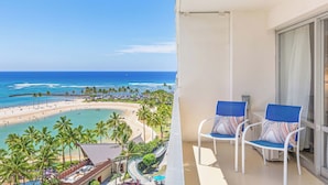 A balcony with two white chairs and a view of a beach with palm trees and a clear blue sky.