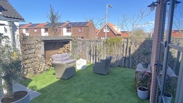 Relax in a serene backyard with a lush lawn, wicker sofas, and a small table, surrounded by potted plants and residential buildings.