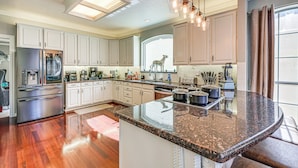 Una cocina amplia con armarios blancos, un refrigerador de acero inoxidable, una isla de encimera de granito e iluminación colgante.