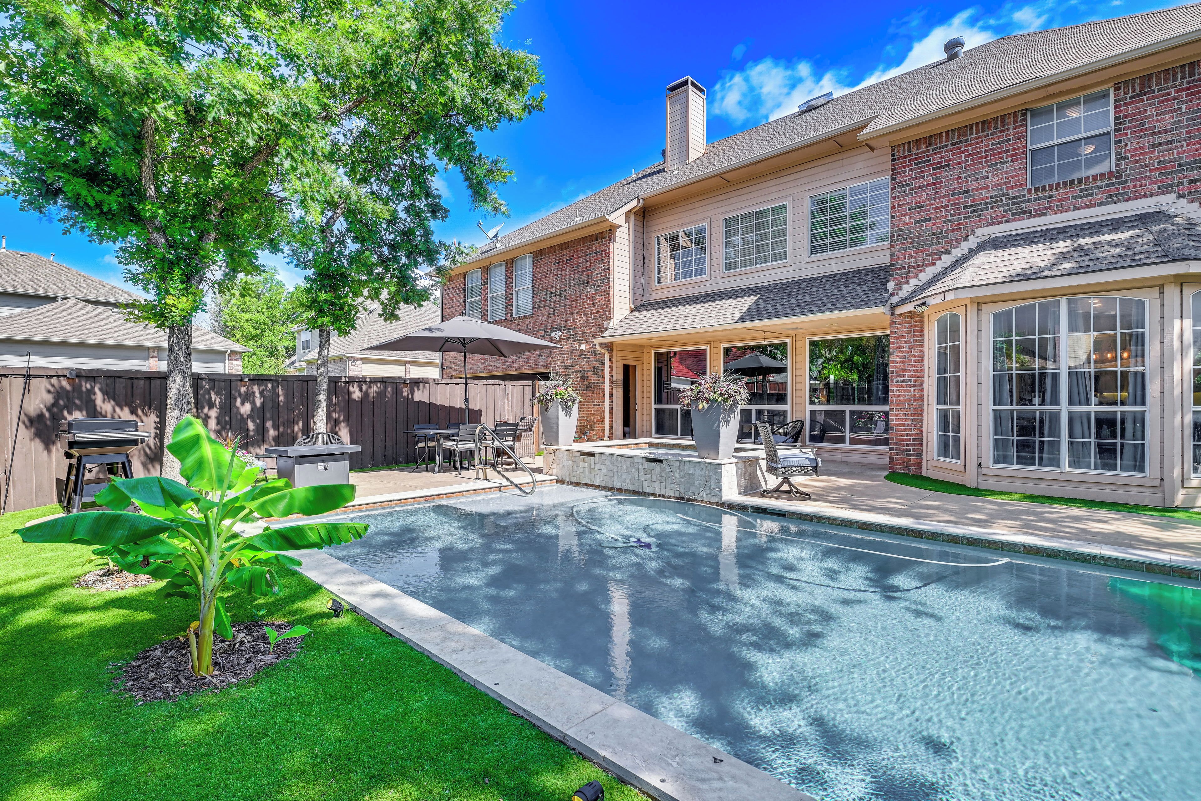 A residential backyard with a swimming pool, a patio area, and a brick house.
