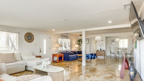 A spacious living area with a large white sofa, a blue sofa, and a coffee table. There is a dining area with a table and chairs in the background.