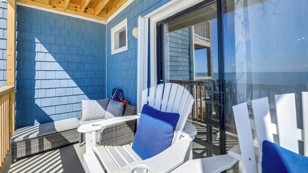 A covered outdoor seating area with Adirondack chairs and a cushioned bench, overlooking a deck and a glass door leading to a balcony. - Oceanfront Gem w/ Views in North Topsail Beach (Sneads Ferry)
