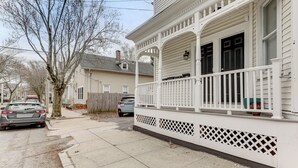 A residential street with a white house featuring a porch with lattice railings and a black door.