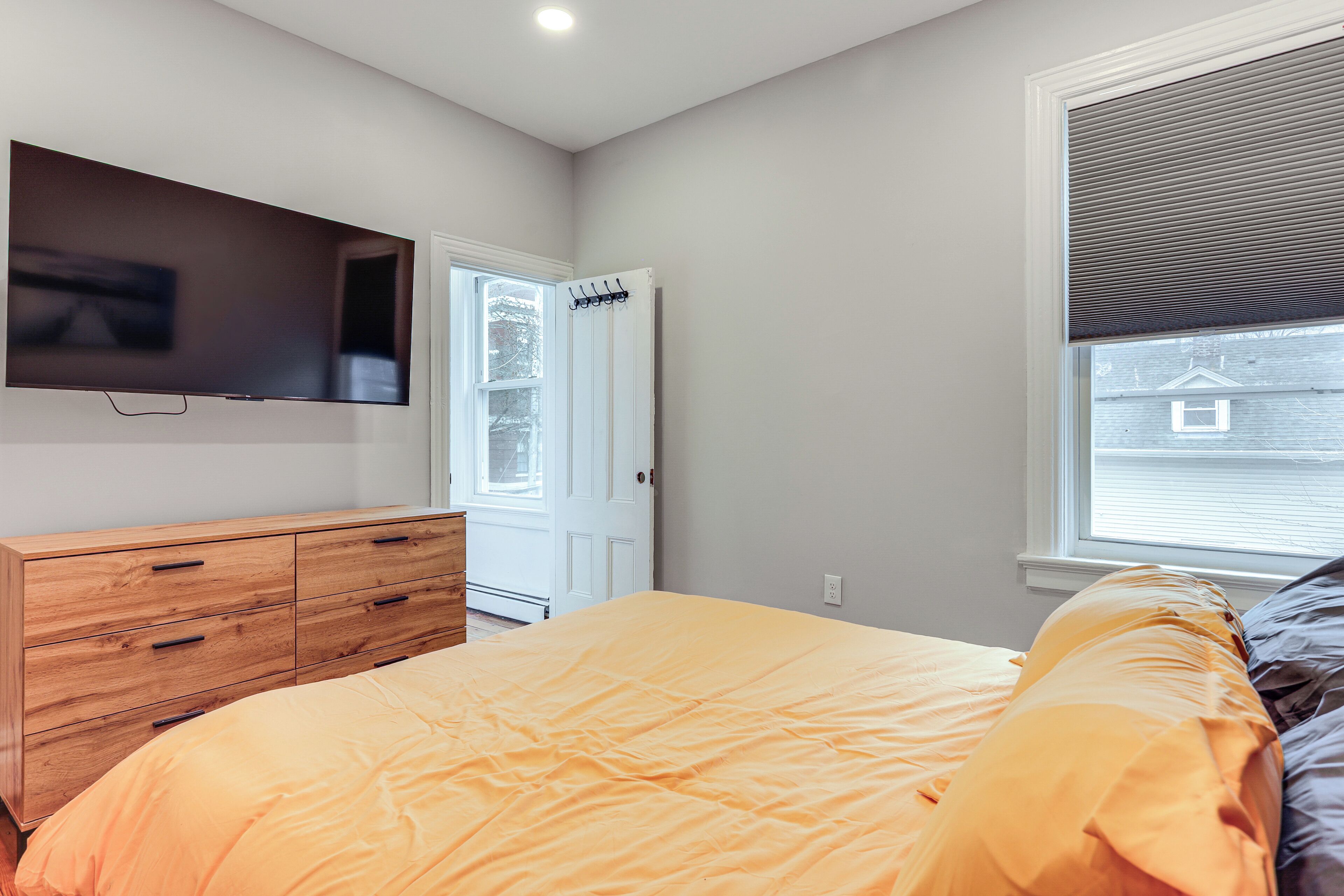 A bedroom with a bed, a wooden dresser, a television mounted on the wall, and a window with blinds.