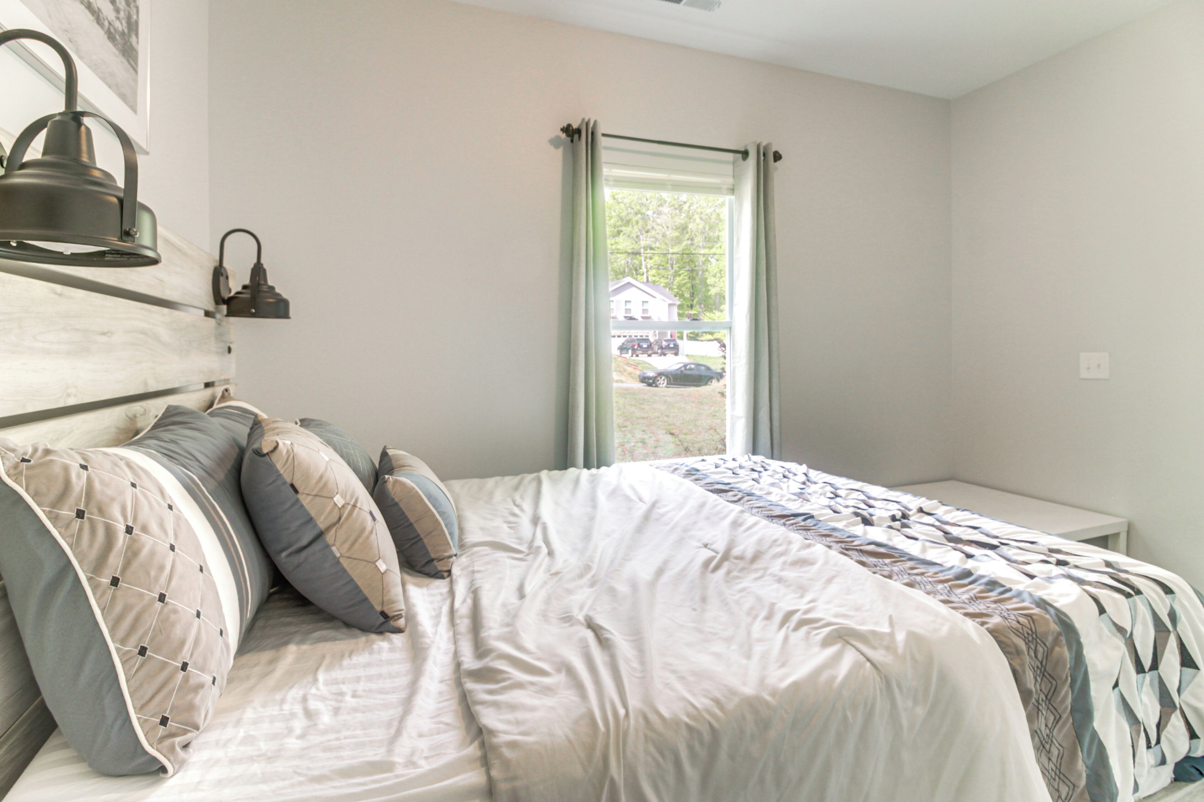 A bedroom with a bed, pillows, a nightstand, a lamp, and a window with curtains.