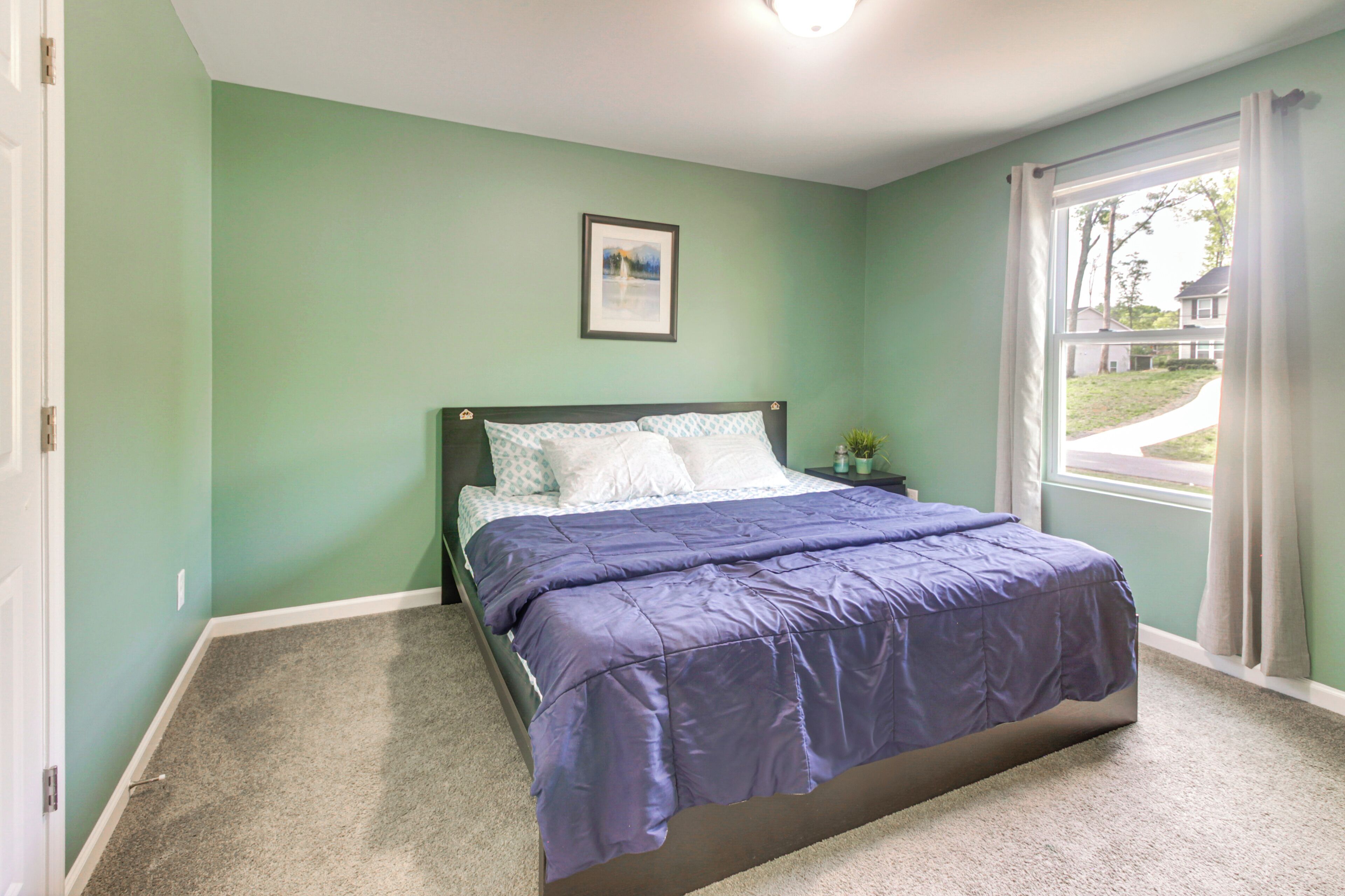 A bedroom with a bed, a nightstand, a framed picture on the wall, and a window with curtains.