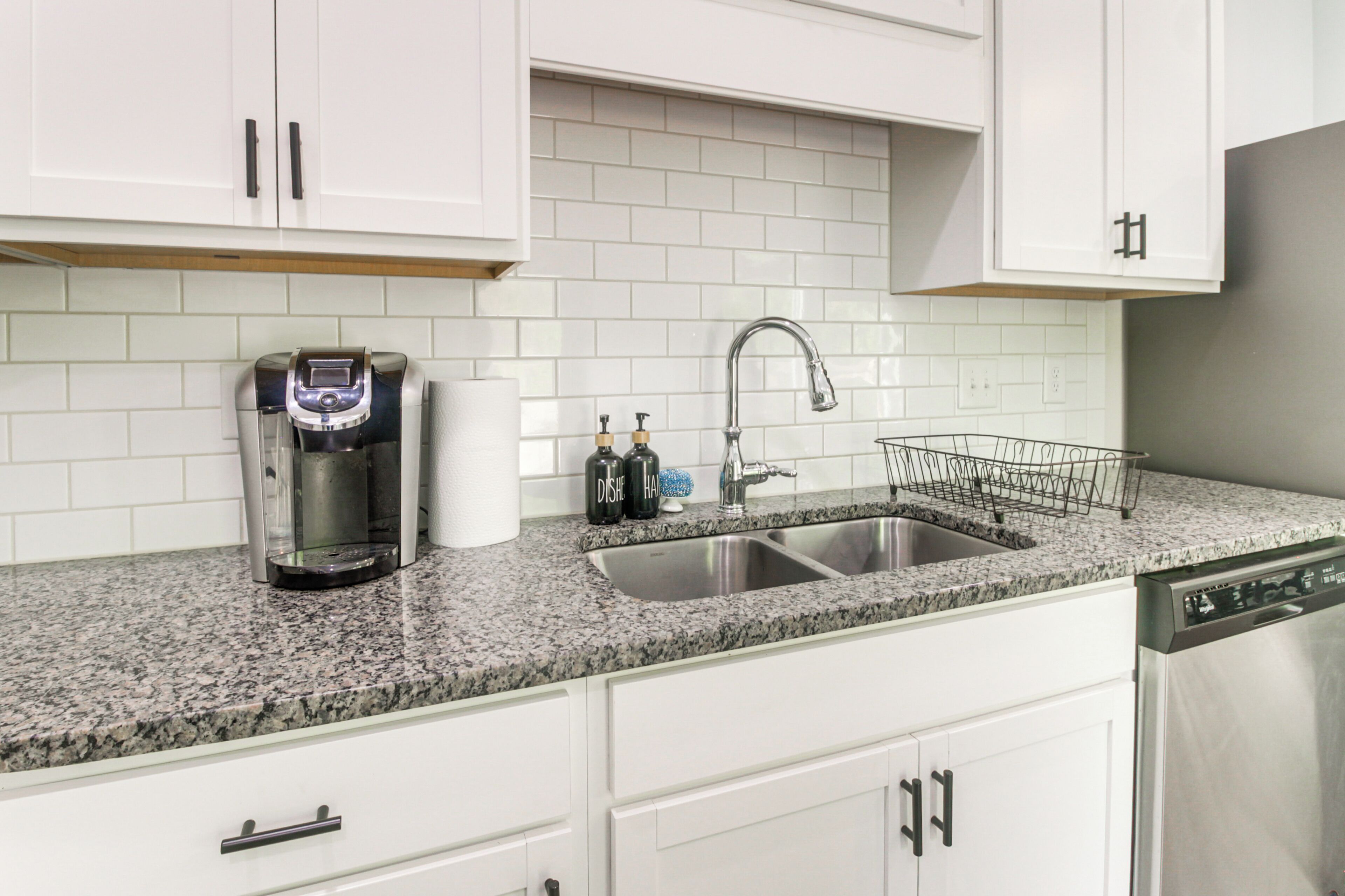 Una cocina con gabinetes blancos, encimera de granito, cafetera, lavabo y escurreplatos.