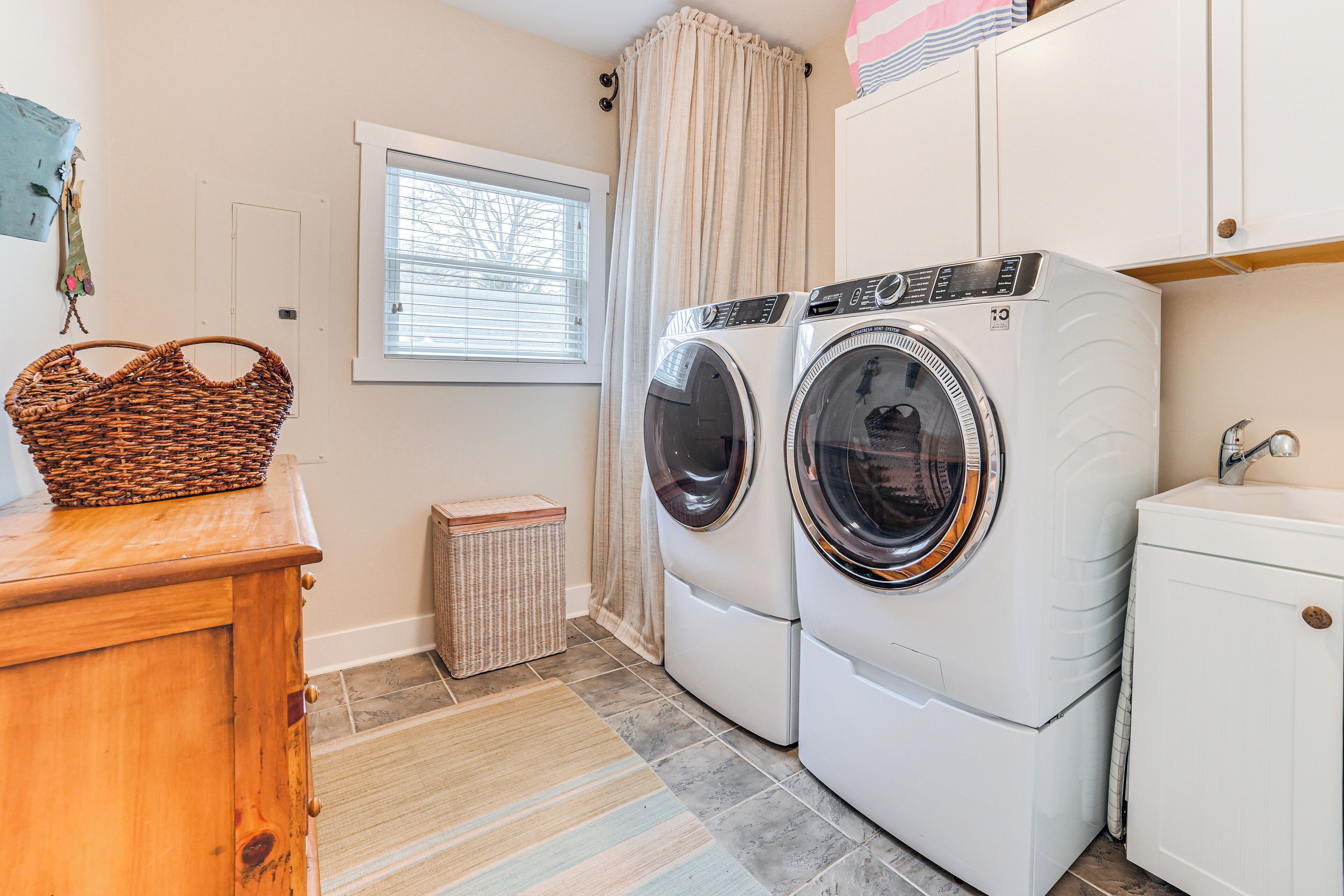 A laundry room with two front-loading washing machines, a wooden cabinet, a wicker basket, a small trash bin, and a window with blinds.