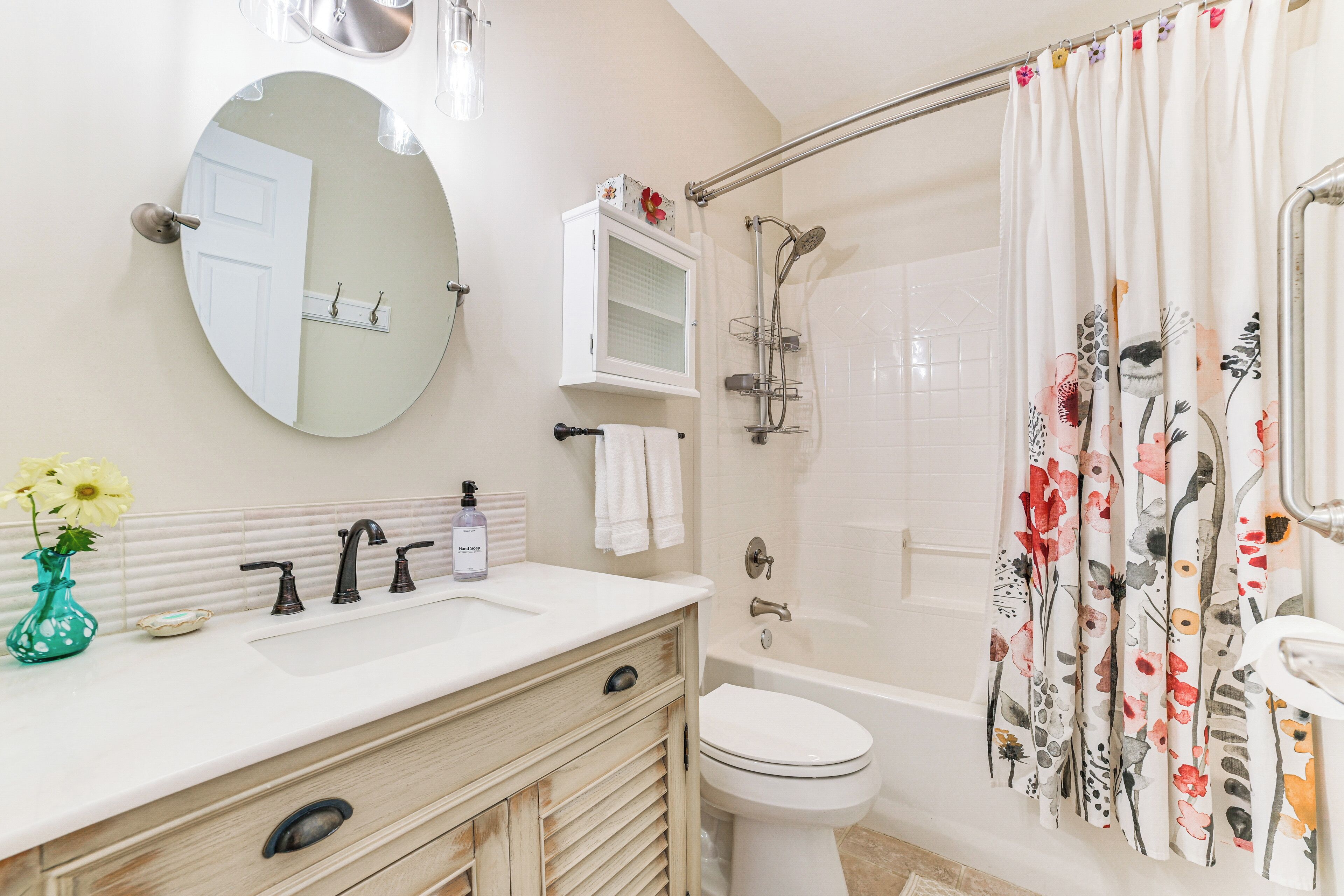 Ein Badezimmer mit rundem Spiegel, einem Waschbecken mit Blumen in einer Vase, einer Toilette und einer Dusche mit floralem Vorhang.