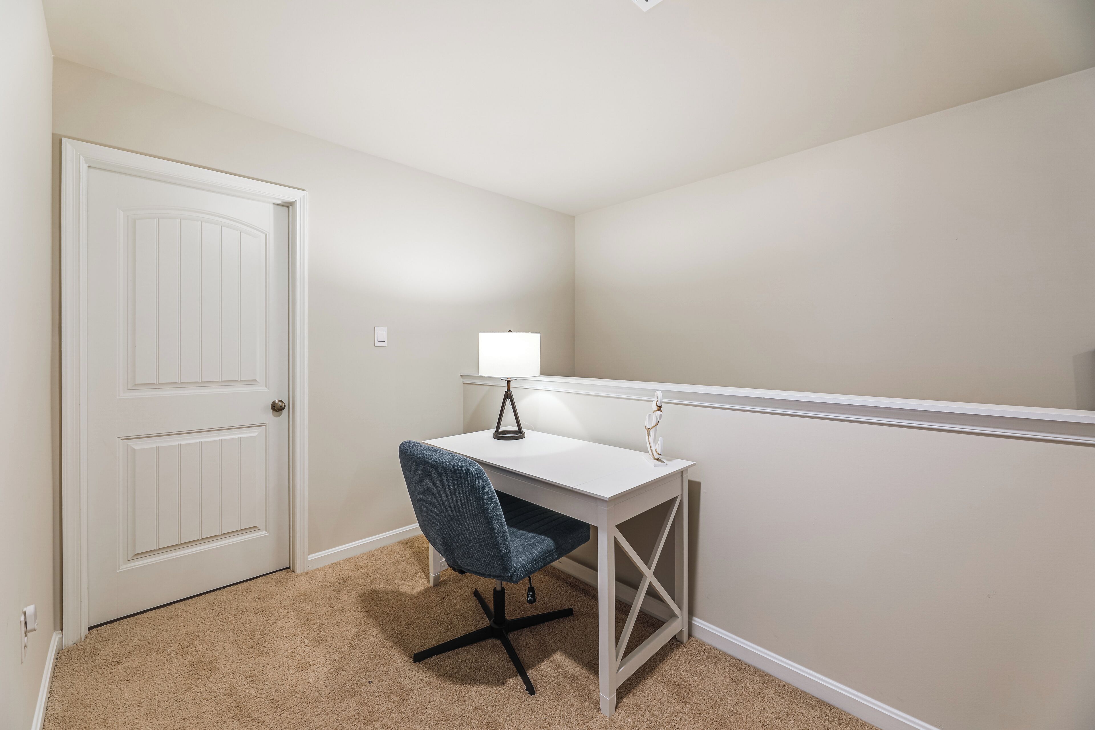 A small, white desk with a blue chair and a lamp in a corner room with beige walls and carpet.