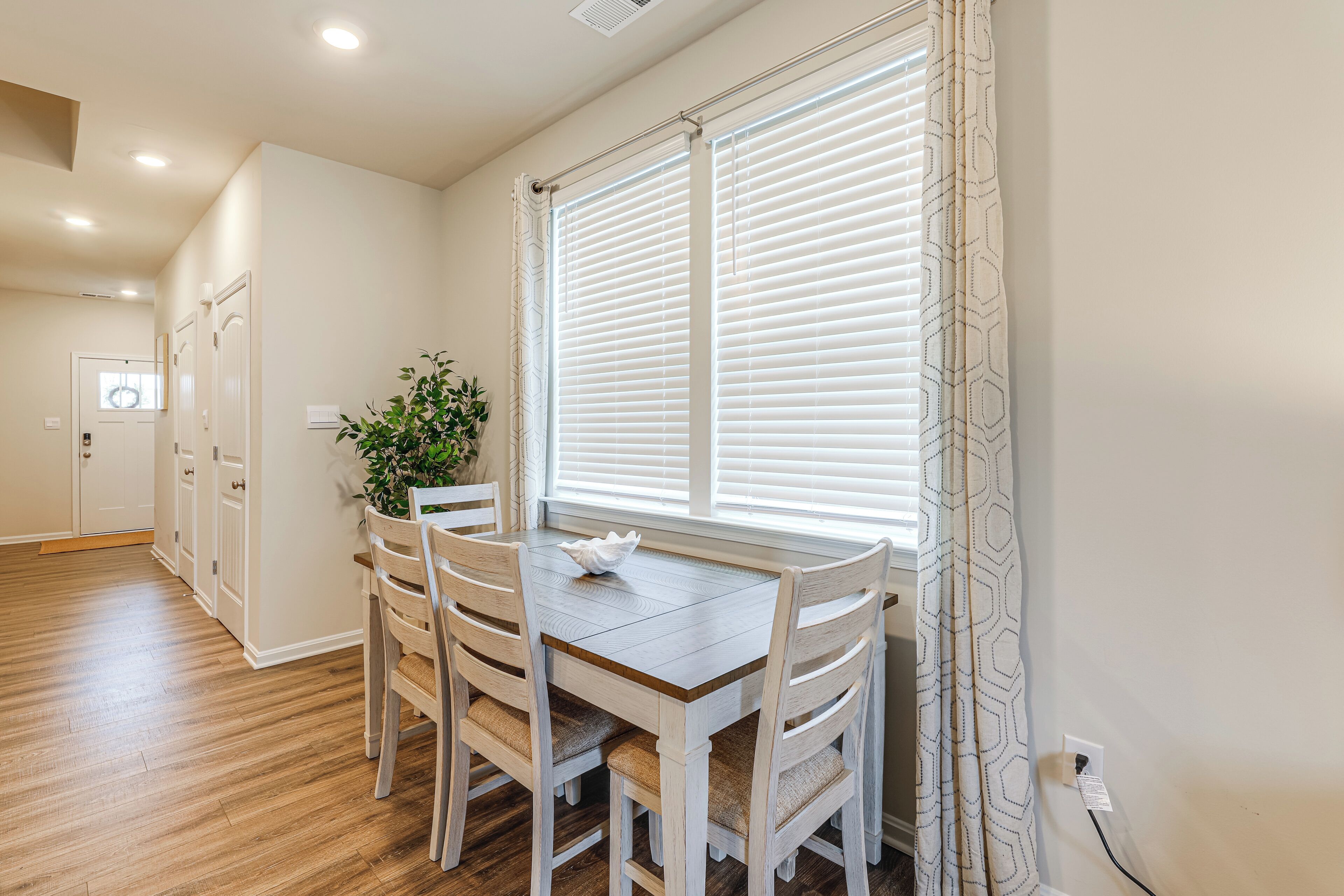 A dining area with a wooden table and chairs, a potted plant, and a window with white blinds.