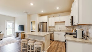 A modern kitchen with white cabinets, a stainless steel refrigerator, a built-in oven, and a granite countertop. There are three stools at the island.