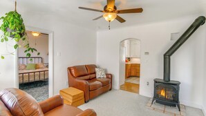 A living room with a leather sofa, a brown armchair, a hanging plant, a ceiling fan, and a wood-burning stove. - Farmhouse w/ Horse Corral Near Wind River Arena (Rigby)