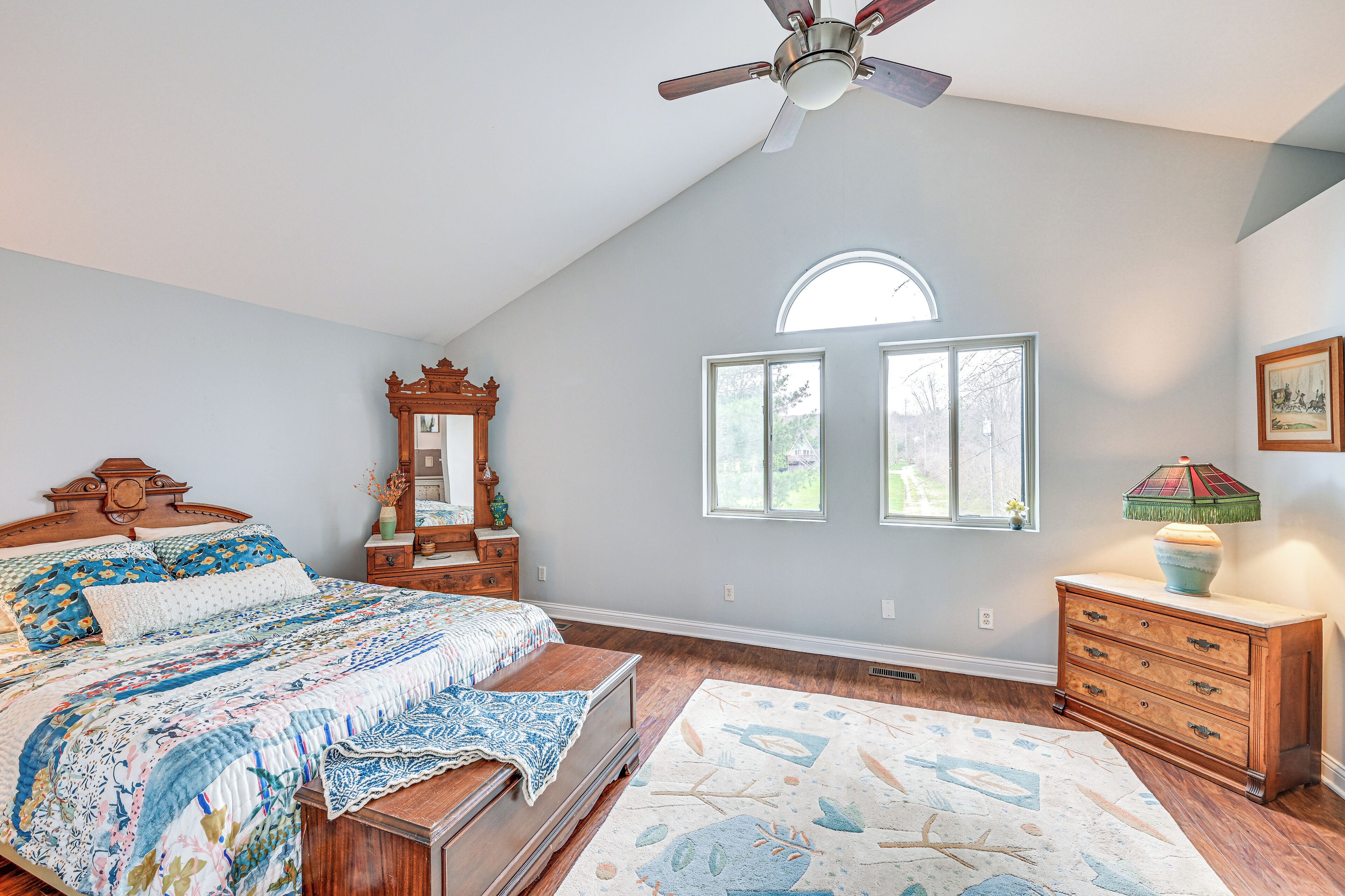 A bedroom with a large bed, a dresser, a mirror, and a ceiling fan. There are two windows and a skylight.