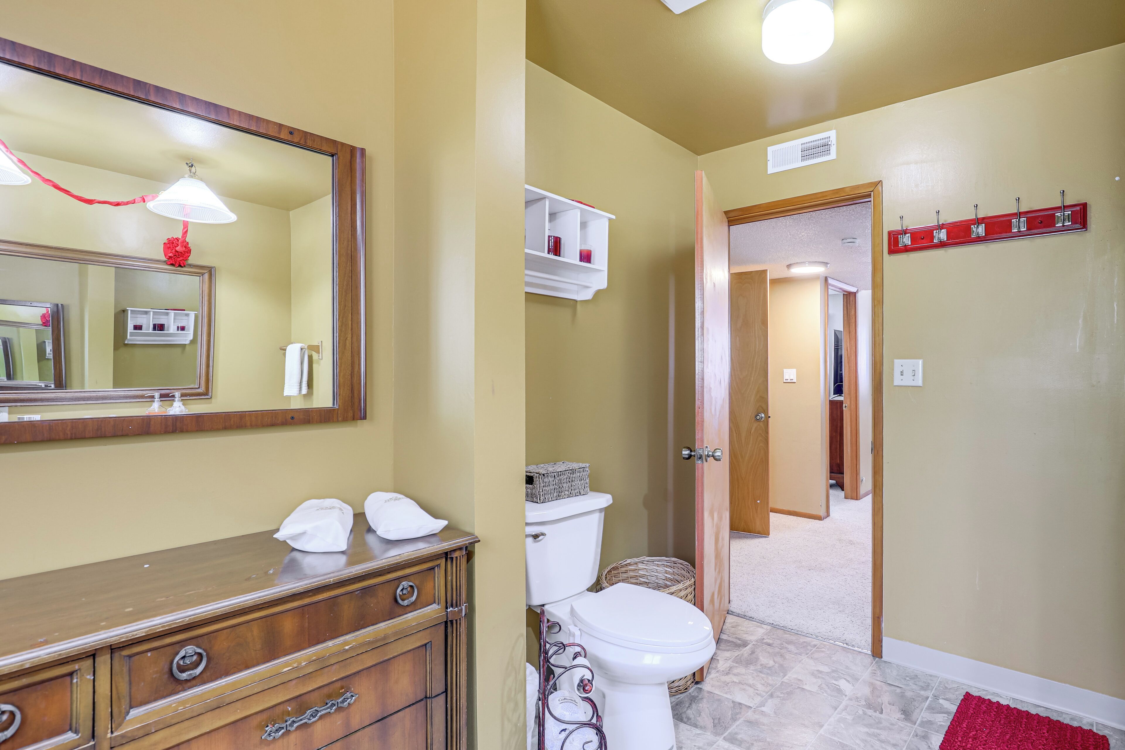 A bathroom with a wooden vanity, a toilet, a mirror, and a red rug.