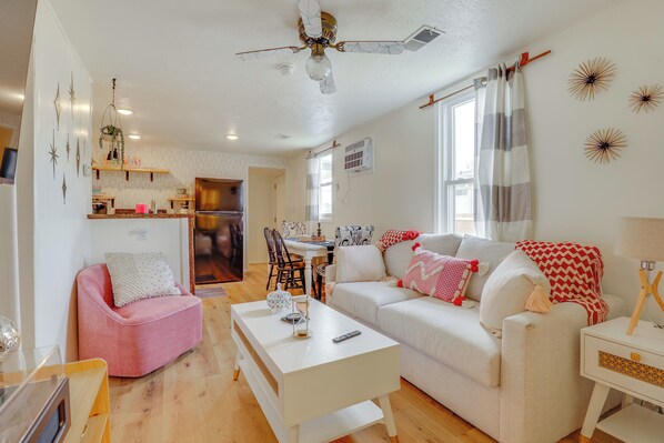 Ein modernes Wohnzimmer mit einem weißen Sofa, einem rosafarbenen Sessel und einem weißen Couchtisch. Es gibt einen Deckenventilator, ein Fenster mit Vorhängen und eine Küchenzeile im Hintergrund.