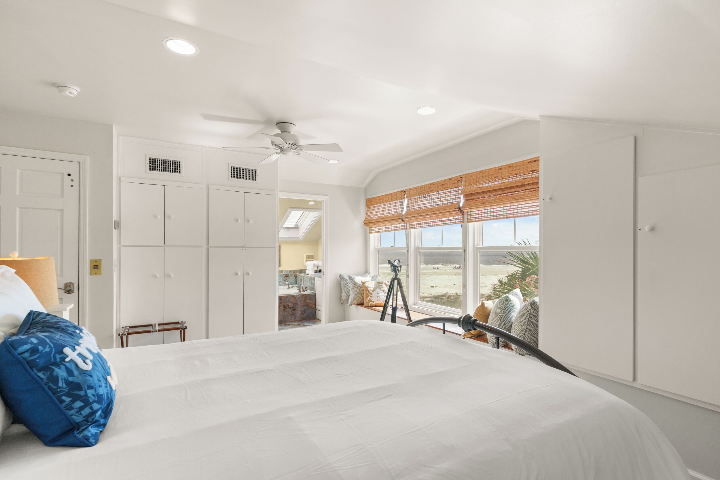 A bedroom with a large bed, a ceiling fan, a window with blinds, and a view of the outdoors.