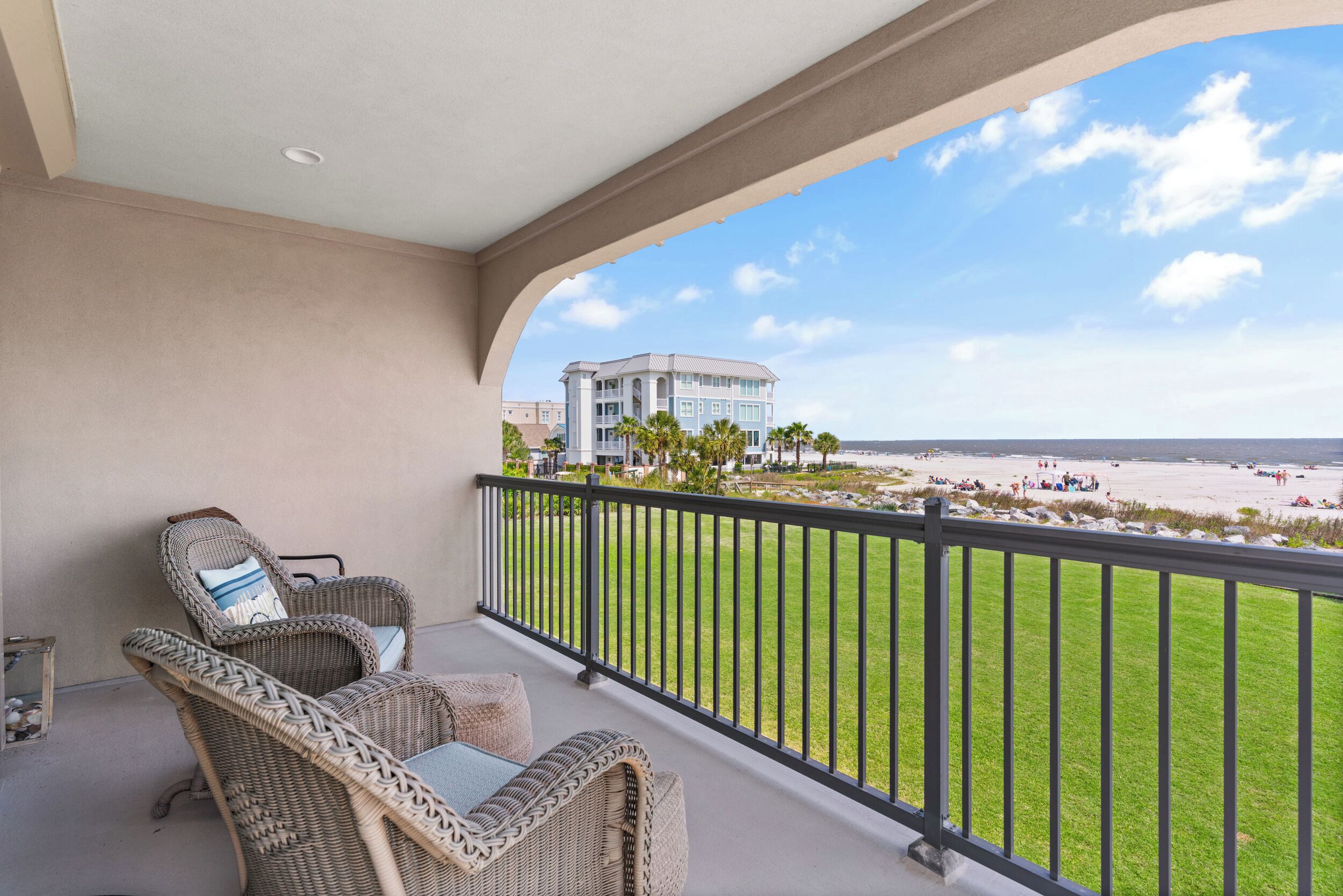 A balcony with wicker furniture, a view of the beach, and a building in the background.