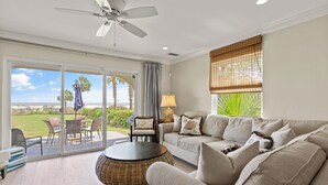 A spacious living room with a sofa, a round wicker table, and a ceiling fan. A large sliding glass door leads to an outdoor patio with a table and chairs. - Unbeatable location with Ocean Views, sleeps 6 (St Simons Island)