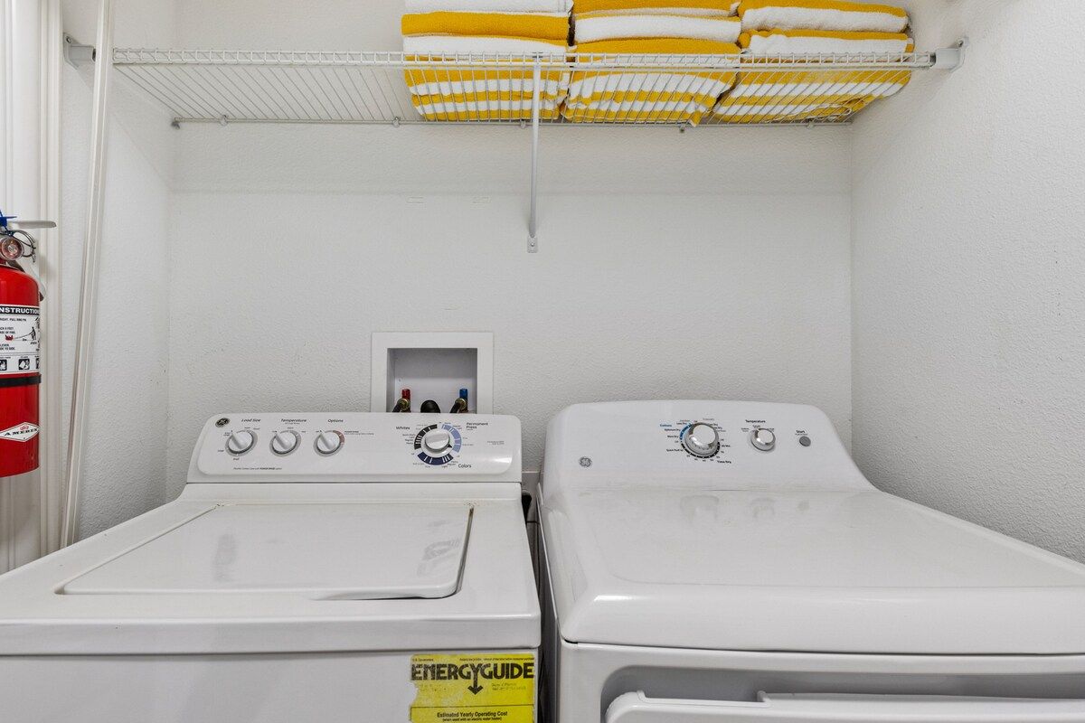 A laundry room with two white front-loading washers and dryers, a fire extinguisher, and a shelf with folded towels.
