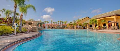 A clear blue pool with a curved edge, surrounded by palm trees and a building with a tiled roof.
