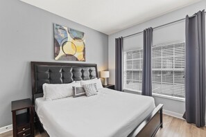 A modern bedroom with a large bed, a headboard, a nightstand, a lamp, and a window with blinds.