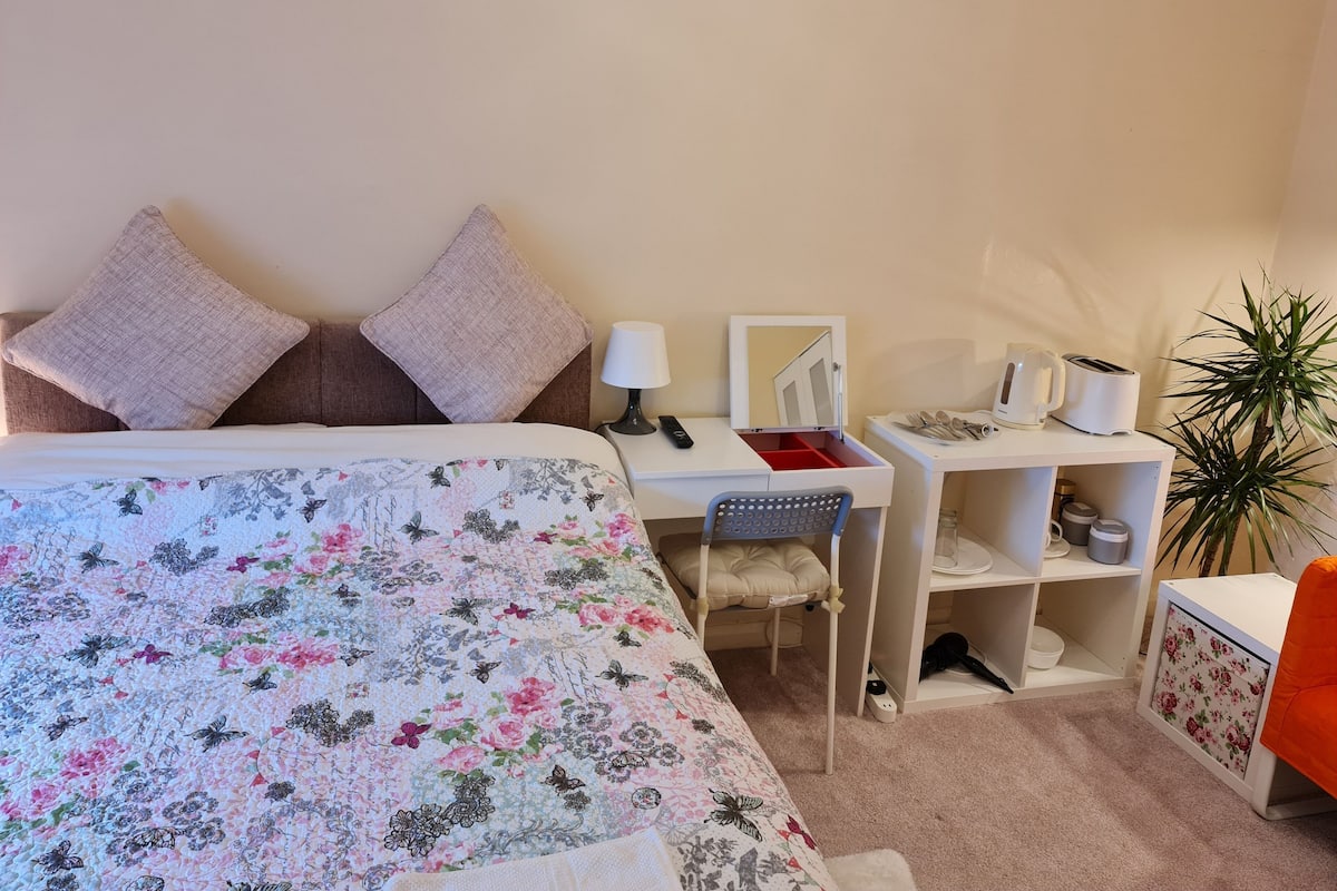 A bedroom with a bed, a desk, a chair, a mirror, a plant, and a shelf with items.