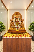 A wooden statue of a deity adorned with garlands and flowers, placed on a marble pedestal in a room with glass walls and potted plants.