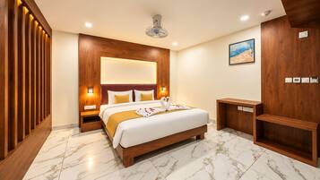 Quarto de hotel com cabeceira de madeira, cama com roupa de cama branca, mesa de cabeceira, um banco de madeira, piso de azulejos de mármore, ventilador de teto e um quadro na parede.