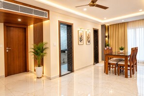 A modern hotel room with a wooden door, a potted plant, a dining area with a table and chairs, and a ceiling fan.