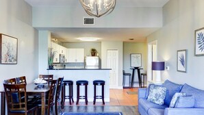 Un salon moderne avec un canapé bleu, une table à manger en bois et des tabourets de bar. On aperçoit une cuisine à l’arrière-plan, avec des meubles blancs et un îlot central.