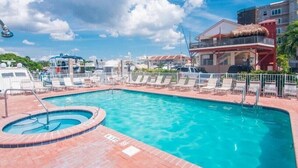 Une piscine entourée de bâtiments, avec des chaises longues et un jacuzzi, sous un ciel dégagé.
